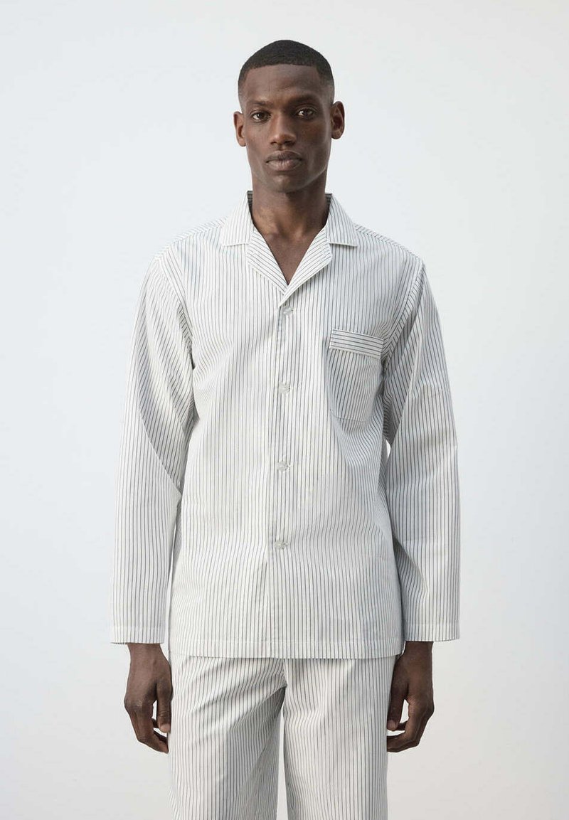 Homme portant un haut de pyjama blanc à manches longues et un pantalon avec fines rayures verticales noires, debout devant un fond clair uni.