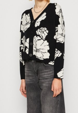 Personne portant un cardigan noir boutonné avec de grands motifs floraux blancs et un jean noir délavé, portant également un collier chaîne en argent.