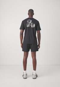 Schwarzes Baumwoll-T-Shirt mit einem Grafikdruck von Hanteln auf dem Rücken. Getragen mit schwarzen Shorts und grauen Sportschuhen.