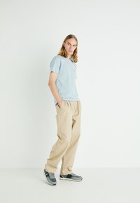 Camiseta de manga corta azul claro, pantalones sueltos beige y zapatillas grises con acentos en marino, de pie contra un fondo blanco liso.