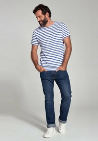 Witte katoenen t-shirt met blauwe horizontale strepen, gecombineerd met donkere denim jeans en witte sneakers. De pasvorm is casual met een ronde hals.