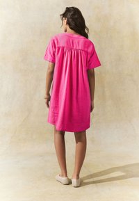 Rozā īss piedurkņu kleita ar savāktu aizmugures dizainu, izgatavota no viegla auduma, kopā ar bēšiem austiem slip-on apaviem.