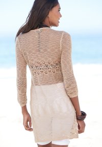 Beige Stricküberwurf mit einem Häkelmuster, Dreiviertelärmeln, offenem Rückendesign und dekorativem Saum mit Diamantakzenten.