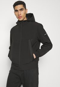 Giacca nera con zip e cappuccio, caratterizzata da un design bicolore, tasche laterali e polsini a coste. Il tessuto appare liscio e resistente.