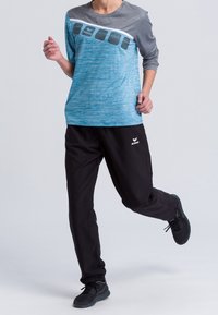 Blaues und graues langärmliges Sportshirt mit strukturiertem Vorderteil und schwarzen Hosen. Mit Logo auf dem Shirt und dem linken Hosenbein, kombiniert mit schwarzen Sneakers.