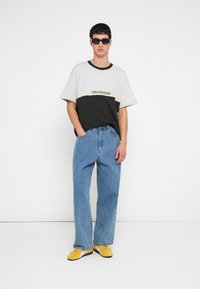 Μαύρο και γκρι μπλουζάκι με μπλοκ χρώματος και λογότυπο, φαρδιά μπλε τζιν τζιν και κίτρινα αθλητικά παπούτσια με μαύρες λεπτομέρειες. Φοράει γυαλιά ηλίου.