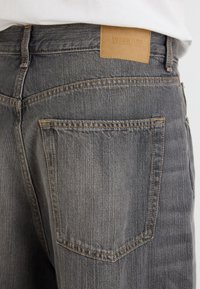 Jean en denim gris avec une finition texturée, doté d'un patch en cuir sur la taille arrière et d'un design classique à cinq poches.