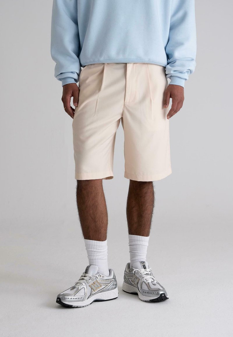 Light beige shorts with pleats, paired with a pale blue sweatshirt and silver athletic shoes. White socks complete the look.