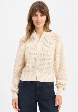 Femme portant un pull zippé en maille couleur crème avec poignets et bas côtelés, accompagné d'un jean en denim foncé sur un fond uni.
