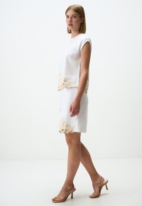 Abito bianco con ricami floreali beige sull'orlo, maniche a campana e una silhouette aderente, abbinato a sandali con tacco nudo.
