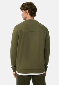 Man in een olijfgroene sweatshirt met lange mouwen en donkere groene broek, gezien van achteren tegen een effen witte achtergrond.