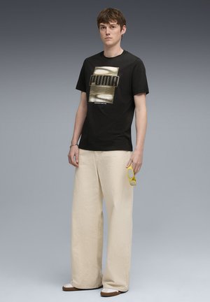 Black cotton t-shirt with "PUMA" graphic in gold, paired with cream wide-leg pants, holding yellow sunglasses, and brown slip-on shoes.