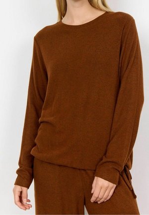 Femme portant un pull en maille marron à manches longues et un pantalon assorti, sur un fond neutre, visible des épaules aux hanches.