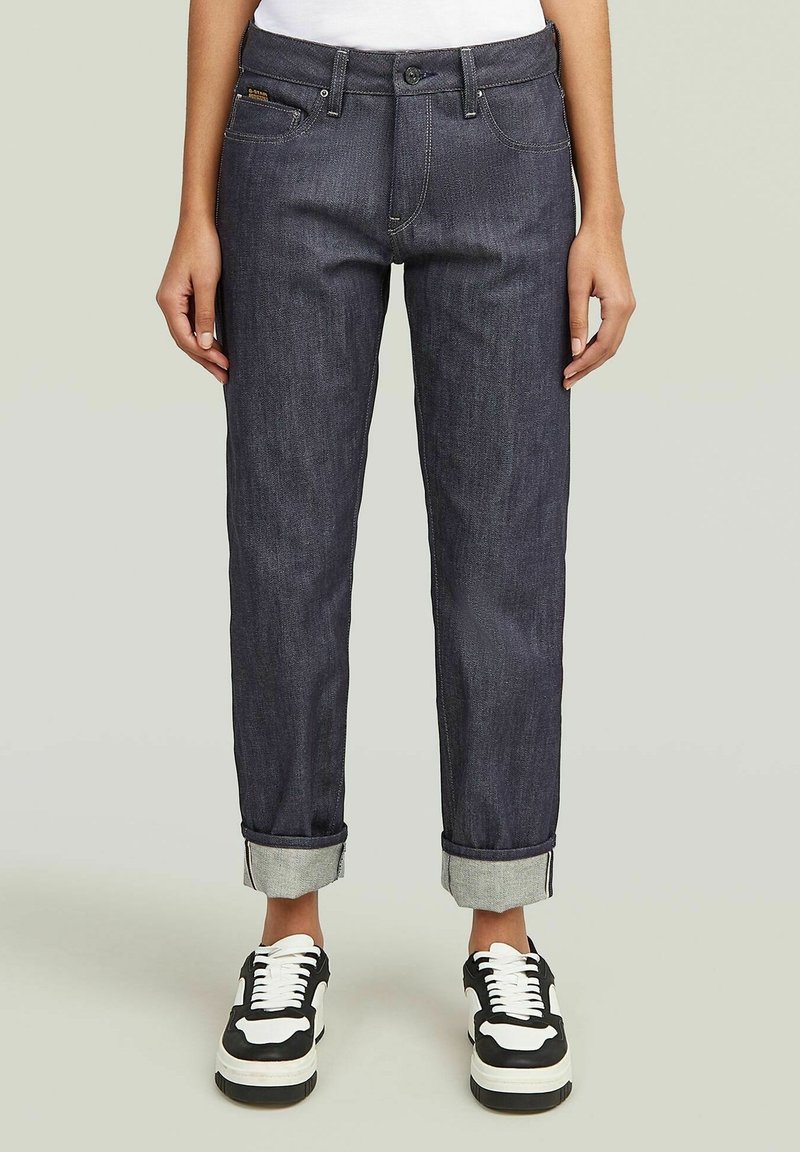 Vaqueros de mezclilla oscura con pierna recta, dobladillos enrollados y costuras contrastantes. Combinados con zapatillas clásicas en blanco y negro.