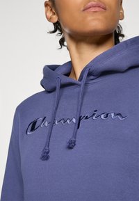 Nærbilde av en person som har på seg en lilla Champion-hettegensere med brodert logo og snor, som viser den nedre delen av ansiktet og nakken.