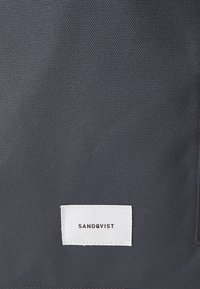 Tessuto testurizzato grigio scuro con un'etichetta bianca che riporta "SANDQVIST" in testo nero. La superficie presenta un motivo sollevato sottile.