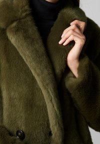 Olivengrøn imiteret pelsfrakke med dobbeltradet design, sorte knapper og en blød, struktureret finish. Nærbillede af krave og hånd, der holder stoffet.