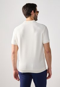 Weißes Polohemd mit kurzen Ärmeln aus strukturiertem Strickstoff, geripptem Kragen und Bündchen, kombiniert mit marineblauen Hosen.
