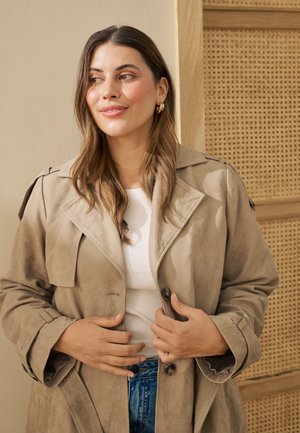 Giovane donna con lunghi capelli castani che indossa orecchini a cerchio dorati, trench beige, top bianco e jeans blu, in piedi davanti a un pannello di rattan.