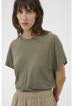 Femme aux cheveux foncés portant un t-shirt à manches courtes vert olive rentré dans un pantalon beige à taille élastique, sur un fond uni.