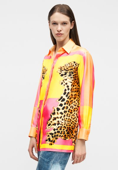 Γυναίκα που φοράει ζωντανό πουκάμισο με κουμπιά, με ροζ, κίτρινα και πορτοκαλί χρώματα σε μπλοκ και μπαλώματα λεοπάρ, συνδυασμένο με μπλε τζιν, σε λευκό φόντο.