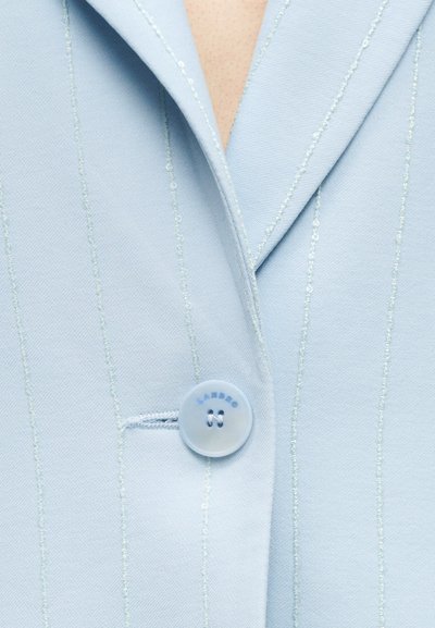 Φόρμα μπλε σακάκι με λεπτές ρίγες, με ένα στρογγυλό κουμπί κουμπωμένο μπροστά, που δείχνει μέρος του γιακά και της γραμμής του λαιμού.
