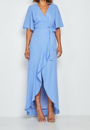 Mujer con un vestido cruzado azul claro con bajo asimétrico con volantes y cintura atada, combinado con sandalias de tacón alto beige con tiras.