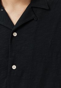 Sort skjorte med krave lavet af tekstureret stof. Har to lyse knapper og en diskret broderet logo på venstre side.