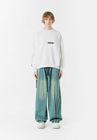 Sudadera blanca de gran tamaño con un bolsillo frontal y texto negro. Pantalones de mezclilla azul claro holgados con un efecto de degradado, combinados con zapatillas blancas gruesas.