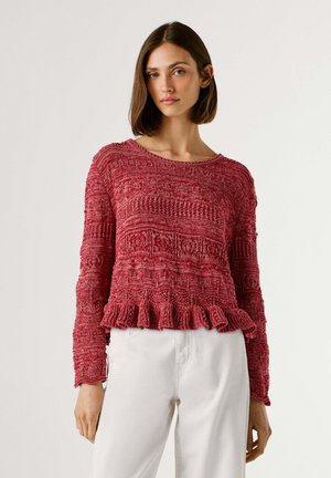 Femme portant un pull en tricot rouge texturé avec un ourlet à volants et un pantalon blanc, debout devant un fond uni.