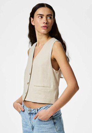 Femme aux longs cheveux foncés portant un gilet beige sans manches et un jean bleu clair, les mains dans les poches, regardant sur le côté.