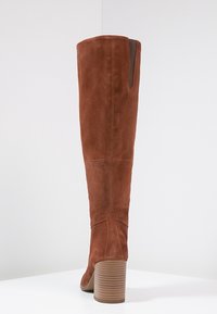 Botte haute jusqu'au genou en daim brun avec talon empilé, panneau élastique latéral visible, photographiée sur une surface blanche avec un fond clair.