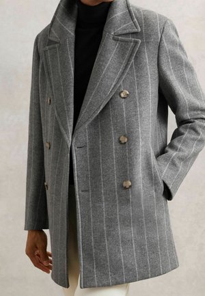 Personne portant un manteau gris croisé à fines rayures avec des boutons marron et un col roulé noir, une main dans la poche, sur un fond uni.
