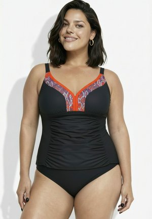 Lächelnde Frau mit schulterlangem dunklem Haar, die einen schwarzen Tankini mit orangefarbener und Schlangenmusterverzierung sowie goldene Creolen trägt.