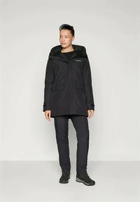 Black waterproof jacket with a hood, featuring two front pockets and a straight cut. Worn with black pants and sturdy black boots.