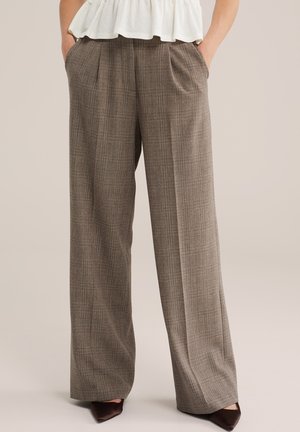 Bruine geruite broek met hoge taille en wijde pijpen met plooien, gedragen met een witte top en bruine puntige schoenen, handen in de zakken.