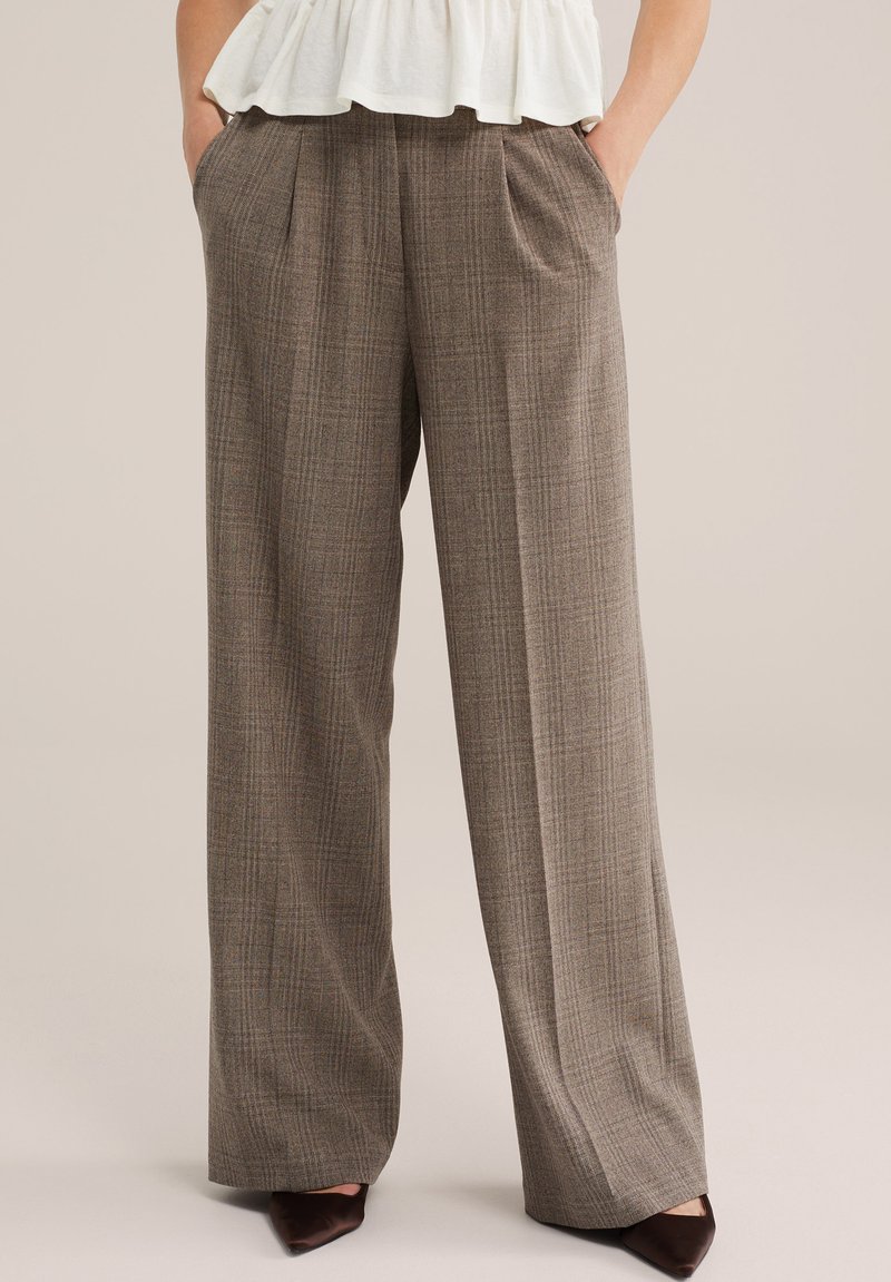 Pantalon à carreaux marron taille haute à jambes larges avec plis, porté avec un haut blanc et des chaussures marron à bout pointu, mains dans les poches.