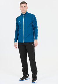 Mand står iført en blå zip-up jakke, sorte træningsbukser og sorte sportsko med blå såler på en hvid baggrund.