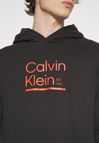 Black hoodie with soft texture, featuring a prominent orange graphic of "Calvin Klein" and "EST. 1968" centered on the chest.