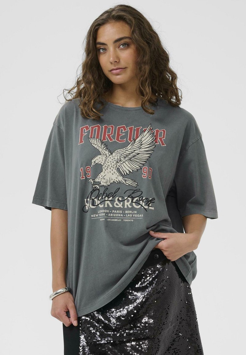 Grå oversized t-shirt med ørner grafik og tekst, med korte ærmer og rund udskæring, båret med en sort paliet nederdel.