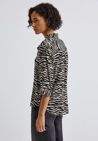 Chemise à rayures zèbre noire et beige, avec des manches trois-quarts, un col et une coupe décontractée. La texture semble lisse et légère.