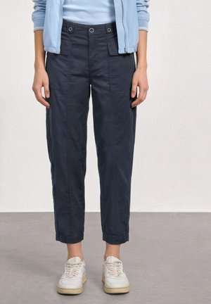 Personne portant un pantalon court bleu marine avec des poches avant, des baskets blanches, un haut bleu clair et une veste bleu clair, debout sur un sol gris.