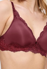 Nainen, jolla on päällä viininpunainen pitsireunuksilla varustettu rintsikka, jossa on sileät kupit ja säädettävät hihnat, keskittyen ylärintaan ja olkapäihin.