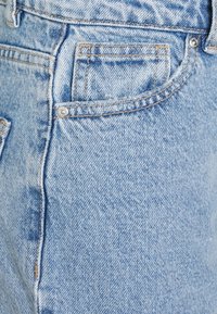 Světle modré džíny z denimu s vybledlým povrchem, mají přední kapsu a viditelné švy. Standardní zapínání na knoflík a zip.