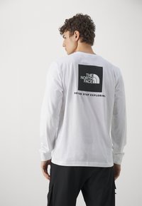 Langærmet hvid t-shirt med et sort firkantet logo på ryggen, der viser teksten "THE NORTH FACE" og "NEVER STOP EXPLORING". Lavet af bomuldsmateriale.