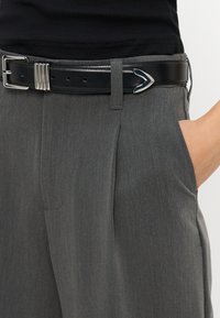 Persona che indossa pantaloni grigi a pieghe con una cintura in pelle nera con fibbia d'argento, mano posata nella tasca destra.