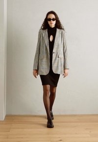 Blazer a quadri in tonalità chiare su un vestito a collo alto nero, abbinato a scarpe nere e occhiali da sole, su uno sfondo minimal.