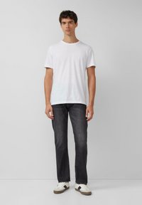 Weißes T-Shirt mit kurzen Ärmeln, kombiniert mit dunklen Jeans und hellen Sneakers, präsentiert an einem Modell vor einem schlichten Hintergrund.