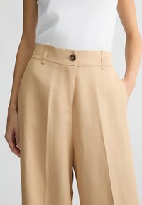 I pantaloni beige a gamba larga presentano un tessuto morbido, una chiusura con bottone, tasche laterali e pieghe sottili per un tocco di stile in più.