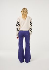 Hellbeiger Pullover mit schwarzen Blumenmustern an den Ärmeln, kombiniert mit lila Schlaghosen. Strukturiertes Strickmaterial mit Puffärmel-Detail.